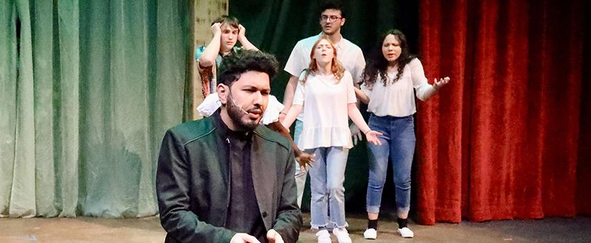 USA Students acting on stage in a play with one male student in front kneeling and 4 students behind him.