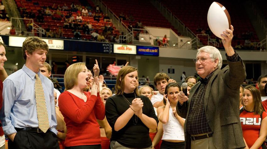 President Moulton holding football in the air in the Mitchell Center with crowd cheering.