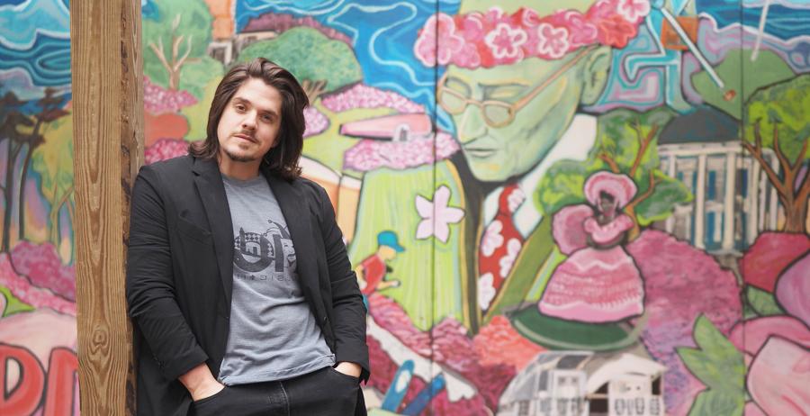 Riley Brenes stands in front of a mural that will be at this weekend’s Festival of Flowers in Cathedral Square. The mural was previously on display in the courtyard at Strickland Youth Center. It features a portrait of Sam Lackland, Mobile scenes and bright pink azaleas. 